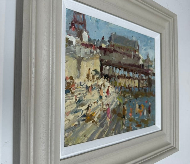 Framed impressionist painting of a seaside promenade or beach scene with figures, buildings, and blue sky, shown in a light wood frame against a white wall.