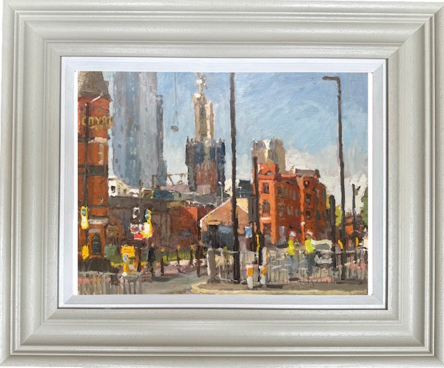 A framed impressionist oil painting of a city street scene, featuring red brick buildings, tall blue-gray skyscrapers, and pedestrians under a light blue sky. The artwork is presented in a wide, ornate white frame with a white mat board.