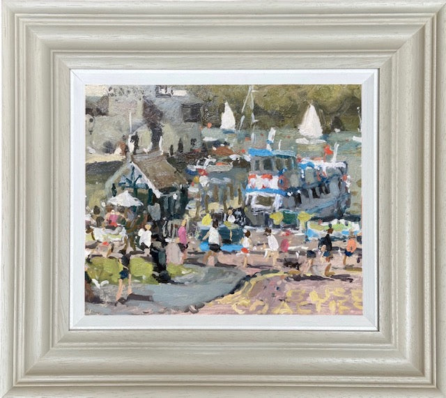 A framed painting depicting a bustling harbor or seaside village scene with boats, figures, and buildings, presented in a wide, light-colored frame with a white mat.