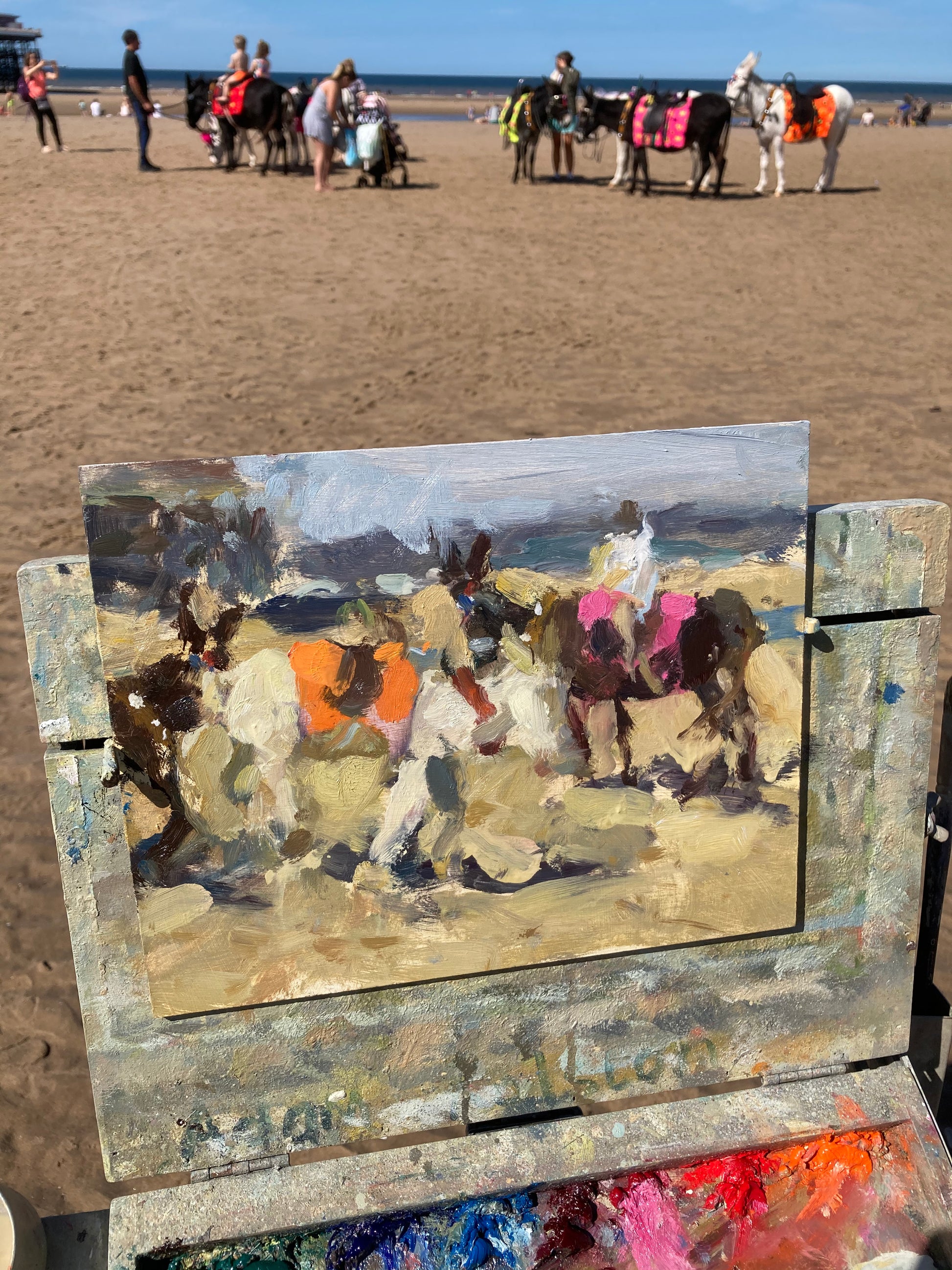 An easel with the painting on it on the beach with the donkeys in the background