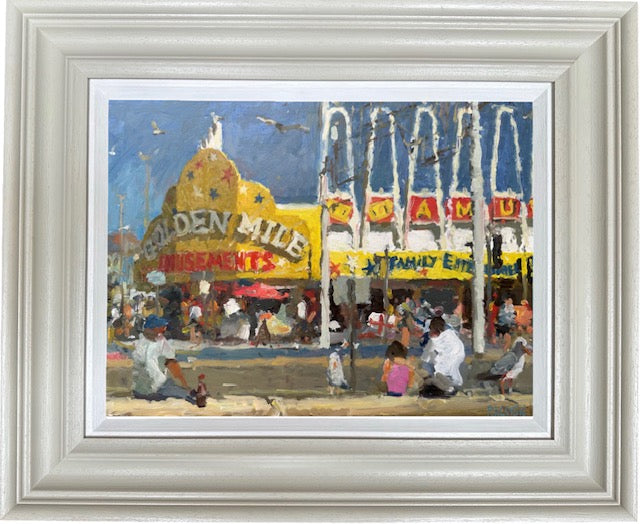A framed art print depicting a colorful, impressionistic scene of a fairground or amusement park with a large yellow 'Golden Mile' building, storefronts, and people in the foreground, set within a wide white frame.
