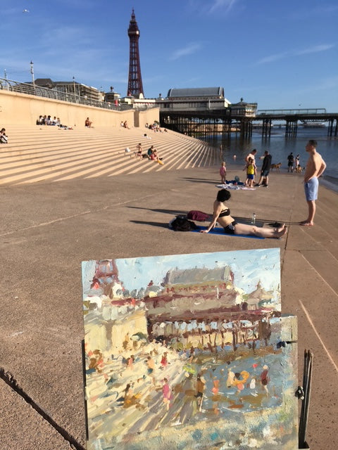 An oil painting of Blackpool's North Pier and Tower at Blackpool beach, with people relaxing on the steps. an outdoor in situ photo of the artist Adam Ralston at work