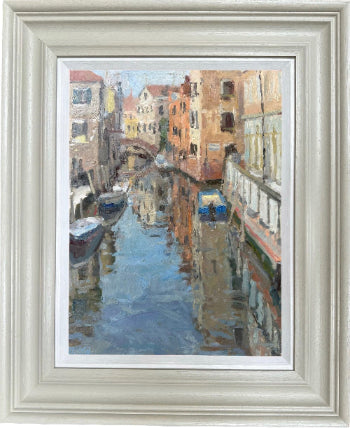Framed impressionist oil painting of a Venice canal scene, featuring warm-toned buildings and blue water reflections, presented in a light gray frame with a white mat.