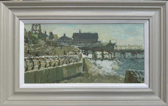 A framed landscape oil painting depicting a coastal pier with buildings and a stone wall, housed in a wide, light gray textured frame.