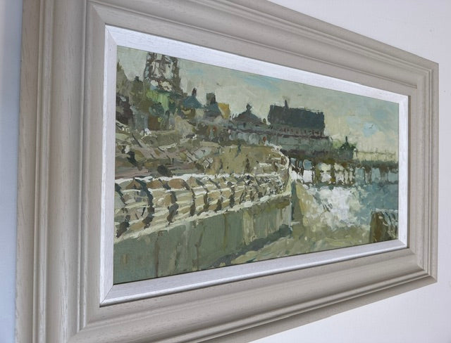 A framed impressionist landscape painting of a seaside town and pier, shown hanging on a white wall. The artwork features muted greens, grays, and beige tones, enclosed in a wide, distressed light gray frame with a white mat.