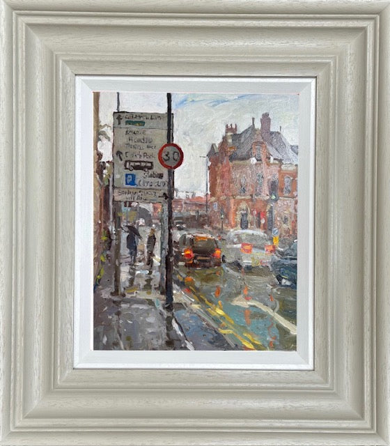 A framed watercolor painting of a rainy city street scene, featuring pedestrians, cars, street signs, and brick buildings. The artwork is presented in a wide, textured light gray wooden frame with a white mat board.