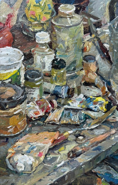 Impressionistic oil painting depicting a cluttered artist's studio table with paint tubes, brushes, jars, and a palette, rendered in textured brushstrokes of gray, beige, brown, yellow, and blue.