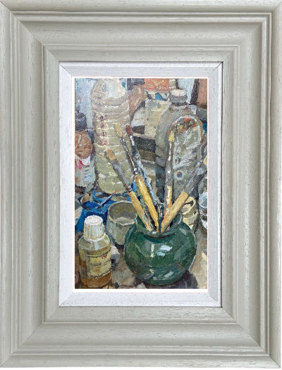A framed still life painting depicting an artist's studio scene with paintbrushes in a green vase, bottles, and tools, set within a wide light gray frame.