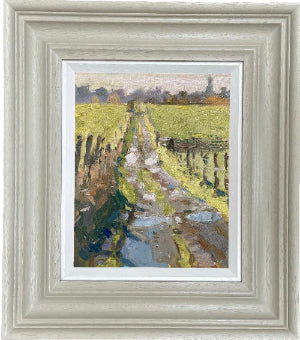 A framed landscape oil painting depicting a stone path leading through a green field, set in a wide, distressed light gray wooden frame with a white mat.