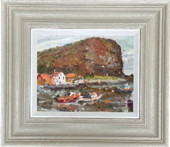 Framed landscape oil painting of a coastal harbor in Staithes with a large reddish-brown rock cliff called Cowbar Nab, small houses with orange roofs, and fishing boats on the water, presented in a light gray wooden frame.