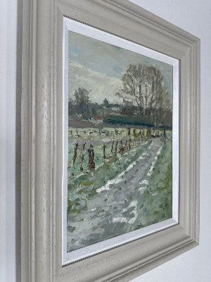 A framed landscape painting of a country path running through a field, shown hanging on a white wall. The artwork features a muted green, gray, and brown palette with a pale sky, and it is set within a white mat and a distressed light gray frame.