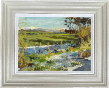 A framed landscape oil painting featuring a green field, a blue stream, and trees under a light sky, housed in a wide white frame with a white mat.