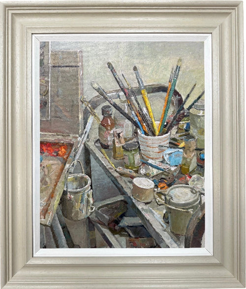 A framed still life painting depicting an artist's studio table cluttered with paintbrushes, jars, bottles, and canvas materials, presented in a light beige frame with a white mat.