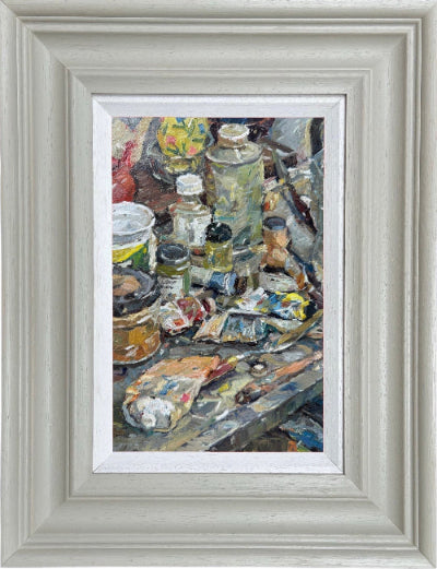 A framed oil painting depicting an artist's studio clutter with paint tubes, brushes, and jars, housed in a wide, light gray textured frame with a white mat.
