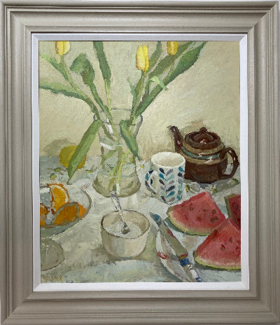 A framed still life painting featuring yellow tulips in a glass vase, a patterned mug, a brown teapot, watermelon slices, and orange slices on a table, presented in a gray frame with a white mat.