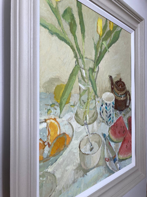 Framed still life painting featuring tulips in a vase, watermelon slices, an orange, a patterned cup, and a teapot on a table, shown in a white frame against a light gray wall.