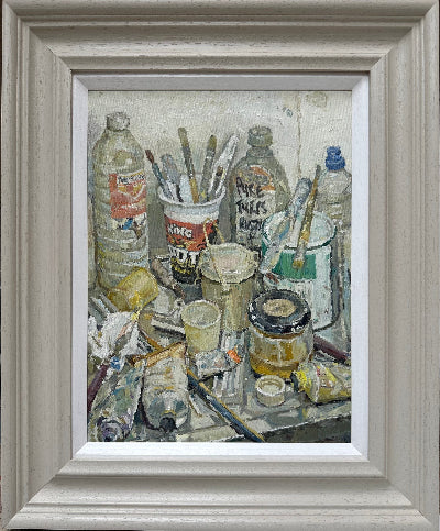 A framed still life oil painting depicting an artist's workshop clutter with paint tubes, brushes, bottles, and jars, housed in a light gray wood frame with a white mat.