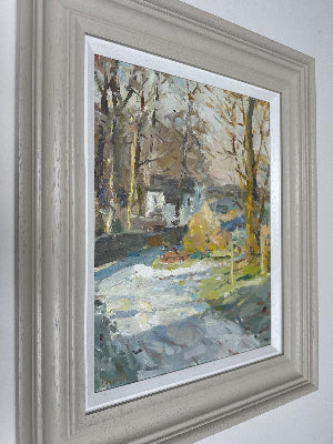 A framed landscape oil painting in Kirkham Wesham featuring a snowy path with trees and a house, presented in a light gray frame with a white mat against a neutral wall.